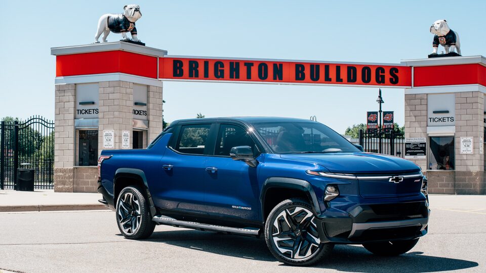 A blue Silverado EV sits in a parking lot in front of a gateway to a stadium. A sign reading Brighton Bulldogs with a bulldog statue on either side is over the gateway.