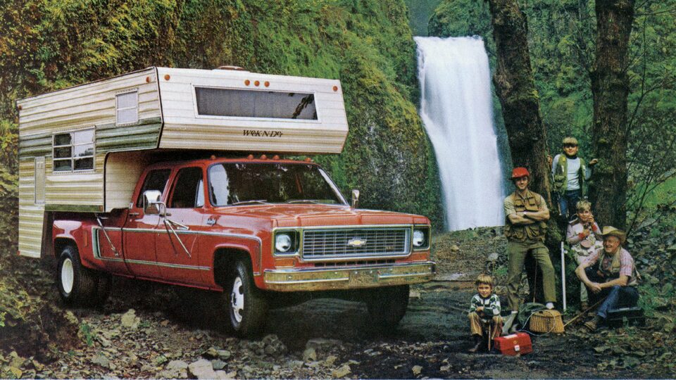 A photo from the original 1973 Chevy truck brochure showing a red C/K Series pickup with a bed camper, sitting in a wooded area near a waterfall. A man and four boys of different ages sit and stand next to the truck.