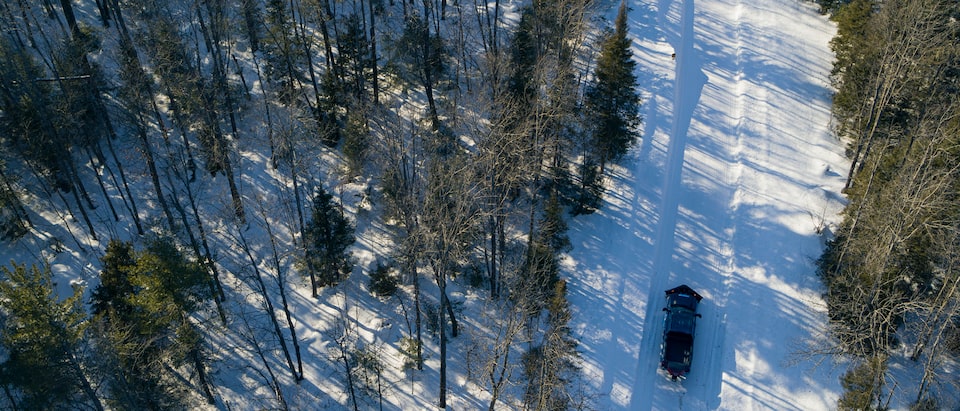 A drone shot of a black Silverado HD with snowplow blade attached driving on a snow-covered road in the middle of a forest.
