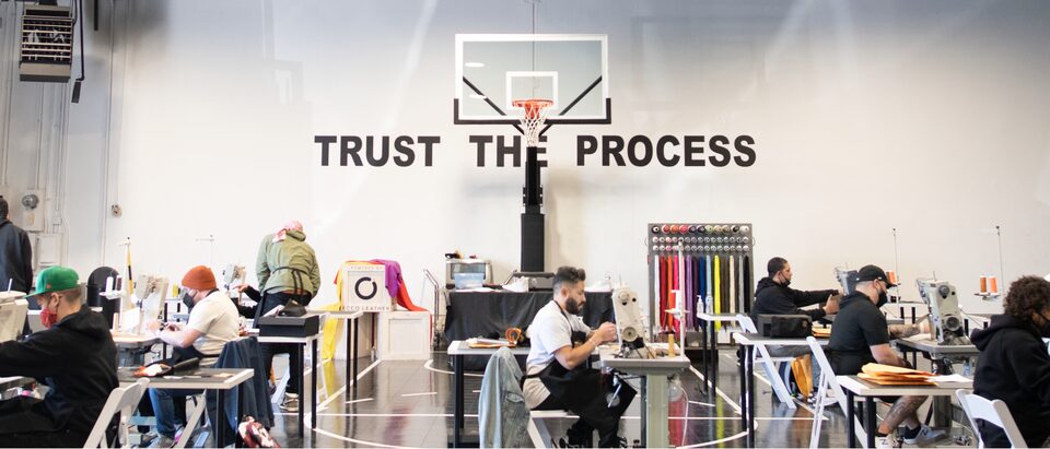 The Sneaker School se instaló en el gimnasio de una escuela, con casi una docena de personas sentadas en mesas con máquinas de coser. Un letrero detrás de una canasta de básquetbol que dice <em>Trust the Process</em> (Confía en el proceso).