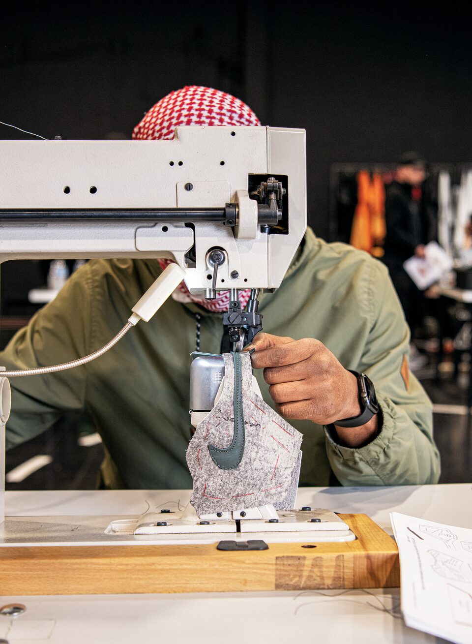 Una persona usando una bandana a cuadros rojos y blancos y una camisa verde trabaja en una máquina de coser industrial, su rostro queda oculto tras la máquina. En la máquina hay una pieza de tela blanca con un logotipo de Nike verde.