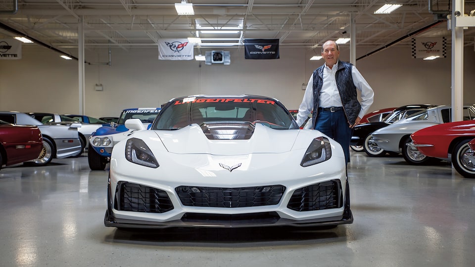 Ken Lingenfelter de pie al lado de un Corvette blanco contemporáneo de su colección, con Corvette antiguos en el fondo.