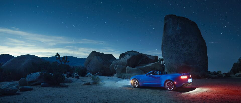 Una vista nocturna de un Camaro Convertible azul con los faros delanteros y traseros encendidos estacionado frente a los afloramientos rocosos en el Parque Nacional Joshua Tree.