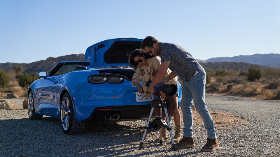 Dos personas montan un telescopio detrás de un Camaro Convertible azul, al lado de un camino de tierra en Joshua Tree National Park.