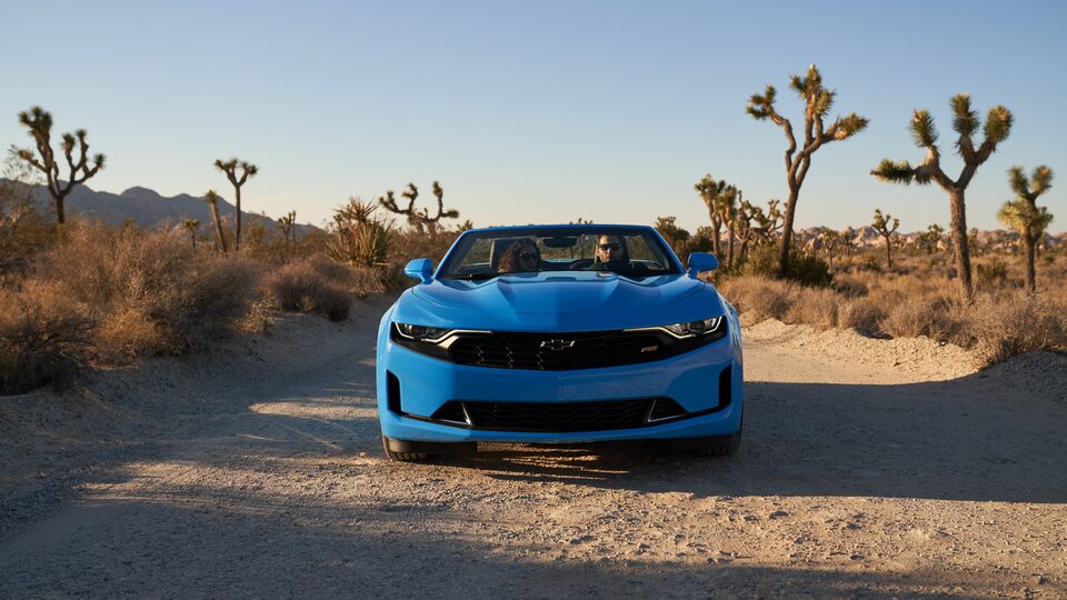 Un Camaro Convertible azul pasa por un camino de tierra en el Parque Nacional Joshua Tree, visto directamente desde el frente del automóvil.