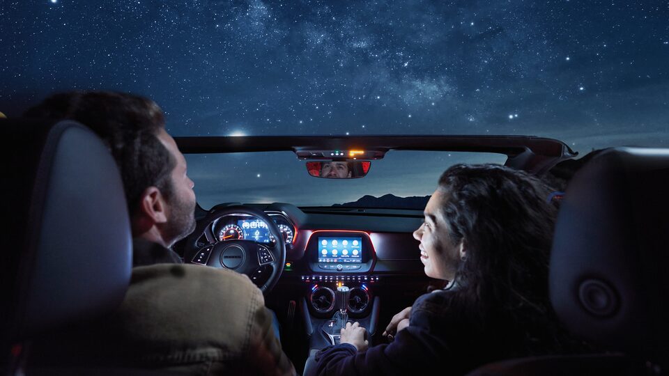 Vista desde el asiento trasero de un Camaro Convertible de noche, dos personas sentadas en los asientos delanteros, iluminadas por las luces del tablero bajo un cielo estrellado y brillante.
