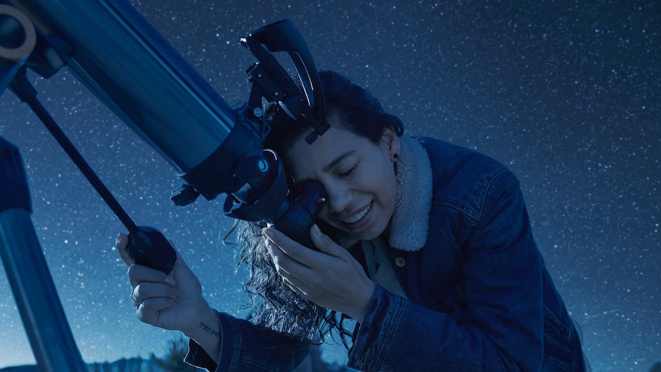 Una persona mirando a través de un telescopio con un cielo azul oscuro y estrellado de fondo.