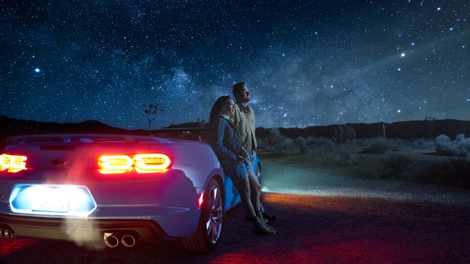Dos personas paradas al lado de un Camaro Convertible en Joshua Tree National Park por la noche, con colinas oscuras y un cielo estrellado de fondo.