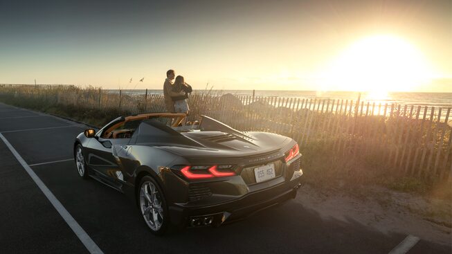 Dos personas viendo el atardecer de pie junto a un Corvette Convertible 2020 en Bronce Zeus Metálico estacionado junto a un área costera en Carolina del Sur.