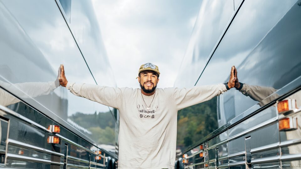 Brad Frey, con una camiseta en la que se lee "All is well,", parado entre dos autobuses de gira estacionados uno al lado del otro y con los brazos extendidos para tocar ambos autobuses.