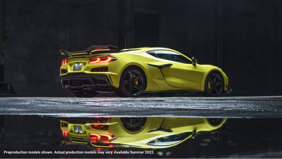 Un Corvette Z06 Amarillo Accelerate Metálico con faros traseros encendidos ubicado en un área oscura, con luces rojas y amarillas borrosas arriba que también se reflejan en el pavimento bajo el auto.