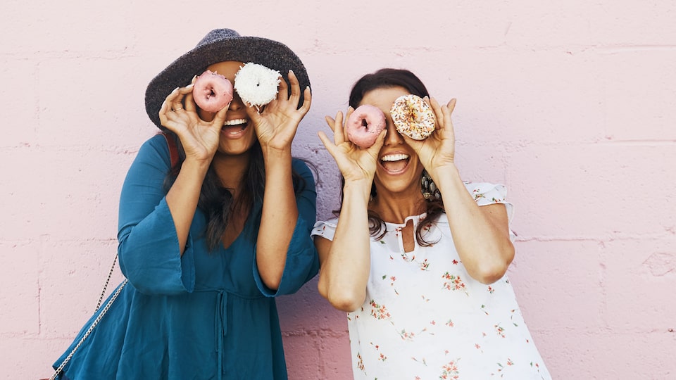 Dos mujeres sonríen sosteniendo donas a la altura de sus ojos.