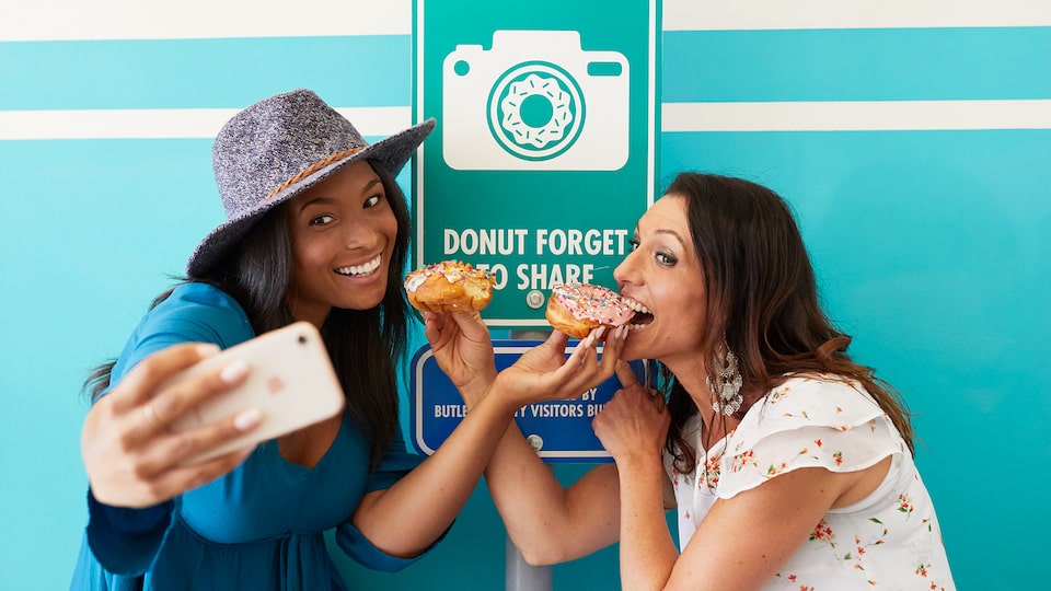 Dos mujeres se toman una selfie posando frente a un cartel que dice "Donut forget to share", haciendo de cuenta que comen donas.