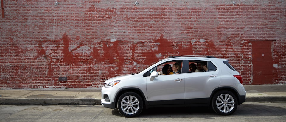 A silver Chevrolet Trax with four occupants sits in front of an old red brick building with faded graffiti on the side.