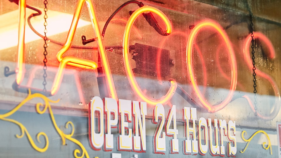 A red neon sign reads “Tacos,” and the window below is painted with the words “Open 24 hours.”