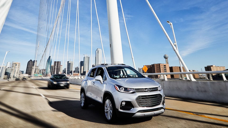 A silver Chevrolet Trax drives across a bridge with the Dallas skyline in the background.
