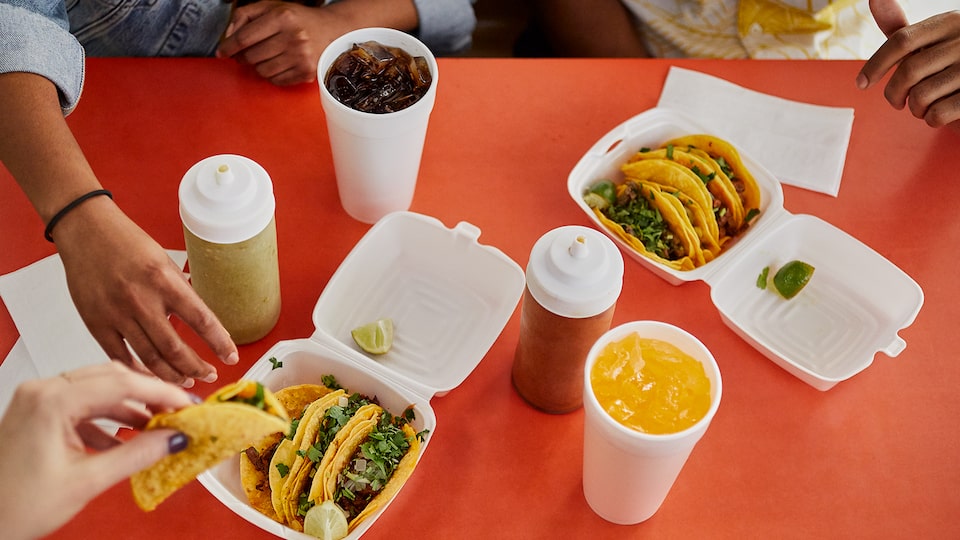 A red tabletop from above with two foam boxes filled with tacos, two drinks in foam cups, and two condiment bottles. Three people sit around the table reaching for the tacos.