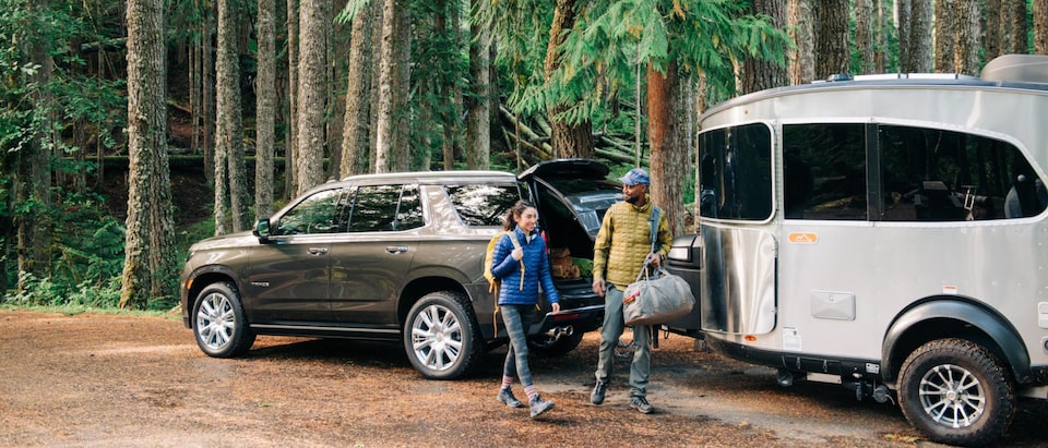 Una pareja joven con equipamiento para acampar, parados al lado de una Chevy Tahoe 2021 con un remolque de viaje Airstream enganchado, estacionada entre árboles altos.
