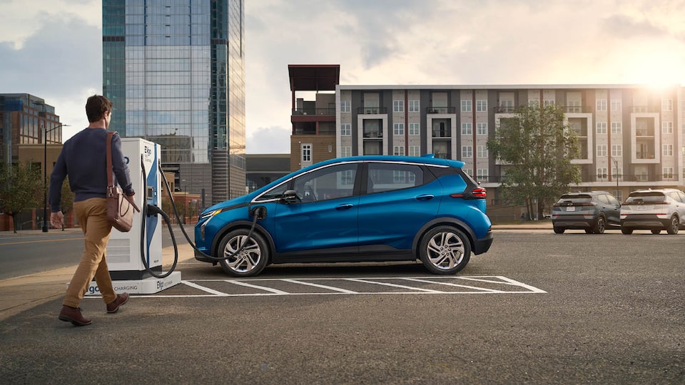 Una persona se acerca a una Bolt EV azul enchufada en una estación de carga pública.