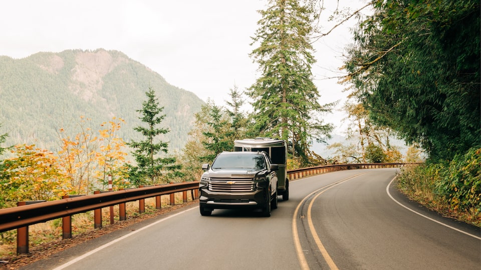 A black 2021 Tahoe pulls an Airstream Basecamp X trailer on a 2-lane highway in a mountainous area.