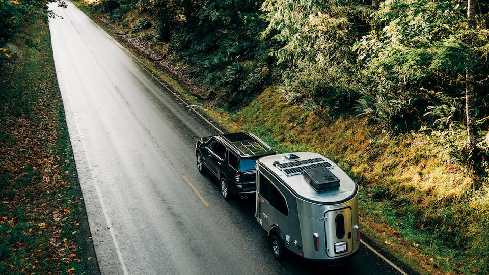 Una Chevy Tahoe negra con un remolque Airstream Basecamp X vista desde arriba andando cuesta abajo por un camino de alquitrán en un bosque.