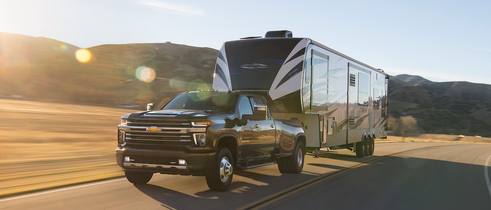 Una camioneta Silverado HD 2020 de color negro transporta un trailer grande por una carretera de dos carriles con colinas en el fondo.