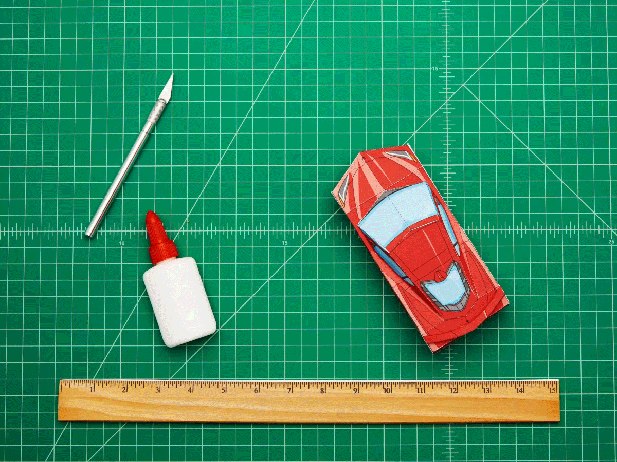 Un gif animado de un modelo Corvette rojo hecho de papel moviéndose por un tablero verde de diseño junto a una navaja para manualidades, un frasco pequeño de pegamento y una regla.