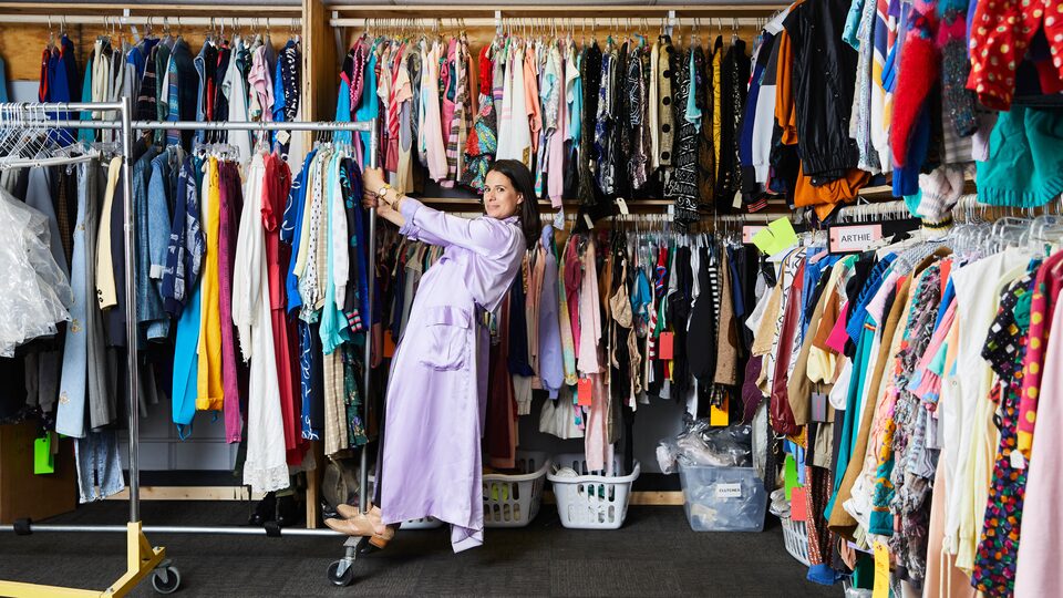 Beth Morgan stands on a portable clothes rack filled with costumes in front of wall racks filled end-to-end with costumes.