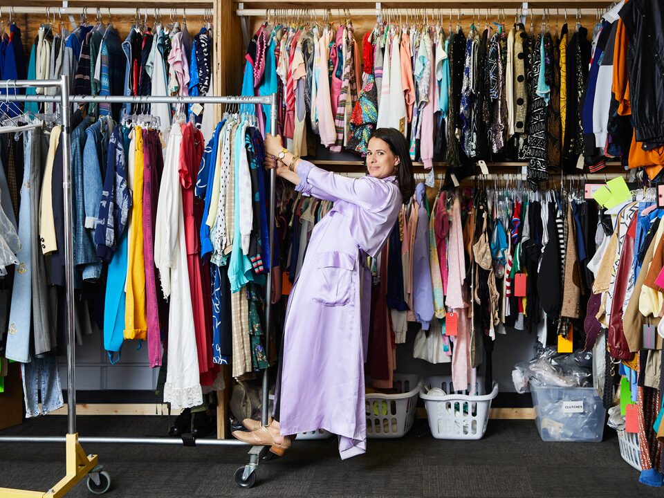 Beth Morgan stands on a portable clothes rack filled with costumes in front of wall racks filled end-to-end with costumes.