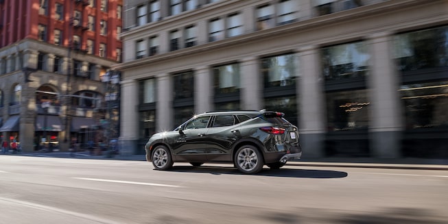 A graphite colored blazer drives past a city building, seen from the driver’s side rear corner.