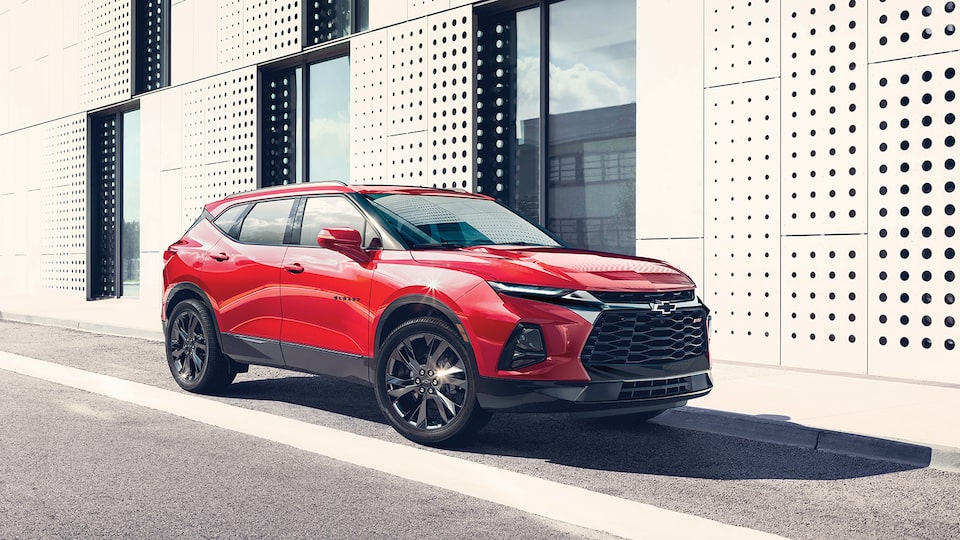 The front and passenger side of a red Blazer RS SUV parked next to a contemporary style building.