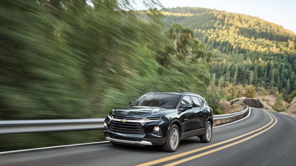 A graphite colored Chevy Blazer drives around a curve on a curvy, tree lined road, seen from the front driver’s side corner.