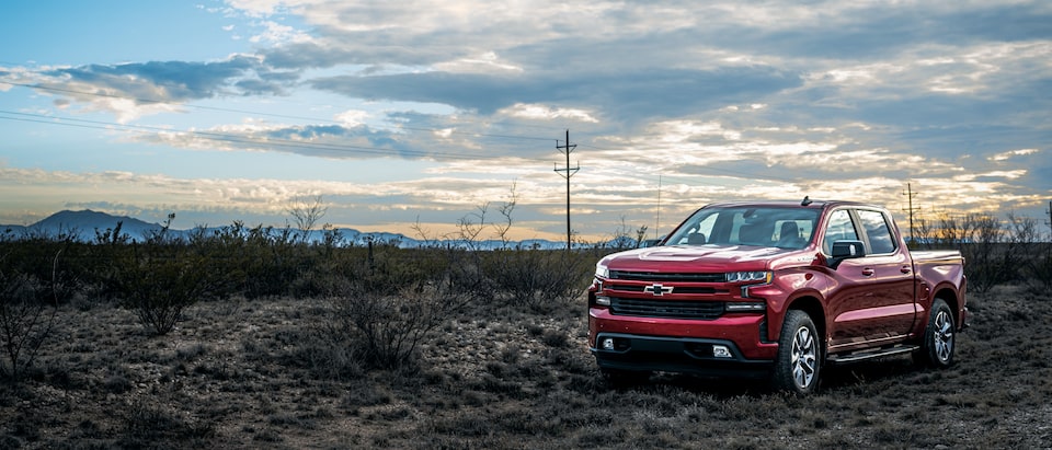 A red Chevy Silverado parked on a Texas roadside surrounded by scrub brush and mountains in the distance, with a partly cloudy sky overhead.