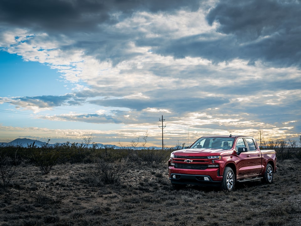 A red Chevy Silverado parked on a Texas roadside surrounded by scrub brush and mountains in the distance, with a partly cloudy sky overhead.