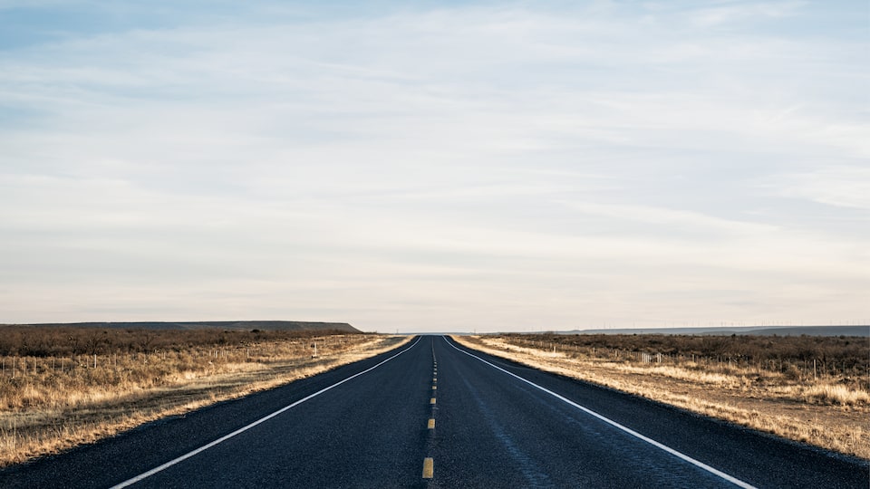A long, straight strip of two-lane highway in Texas disappears at the horizon, lined on either side by scrub brush and a vast cloudy sky overhead.