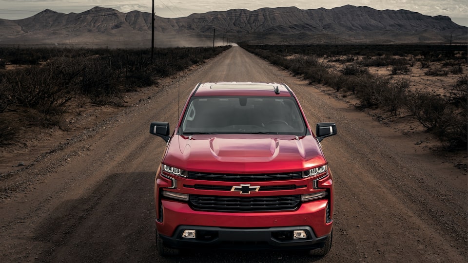 A red Chevy Silverado drives down a lonely dirt road with low mountains in the background and scrub brush on either side.