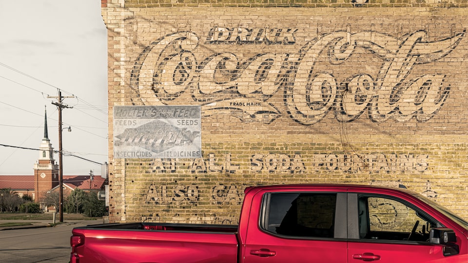 A red Chevy Silverado is parked in front of a faded, hand-painted Coca-Cola advertisement on the side of a tan brick building. A church steeple can be seen in the distance.