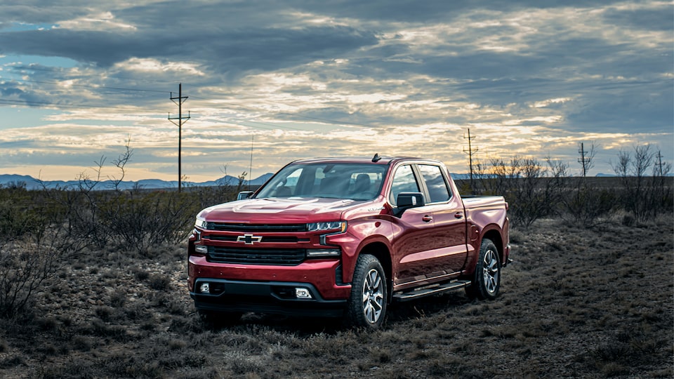 A red Chevy Silverado parked on a Texas roadside surrounded by scrub brush and mountains in the distance, with a partly cloudy sky overhead.