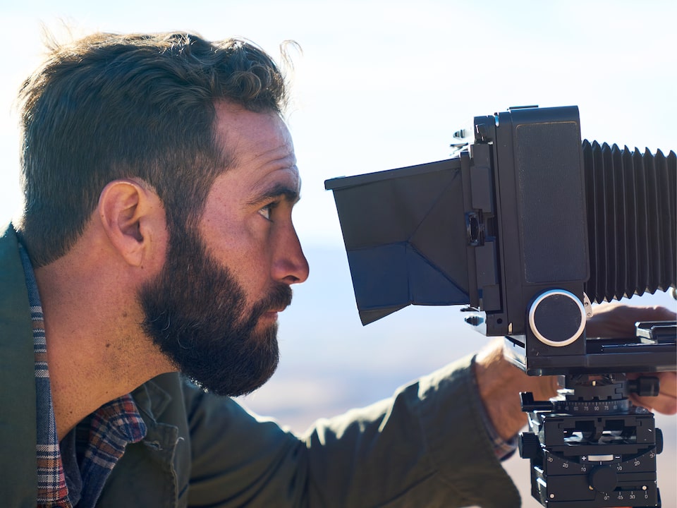 Un hombre con barba mira a través del lente de una cámara de gran formato en frente de un fondo fuera de foco del desierto alto de Arizona.