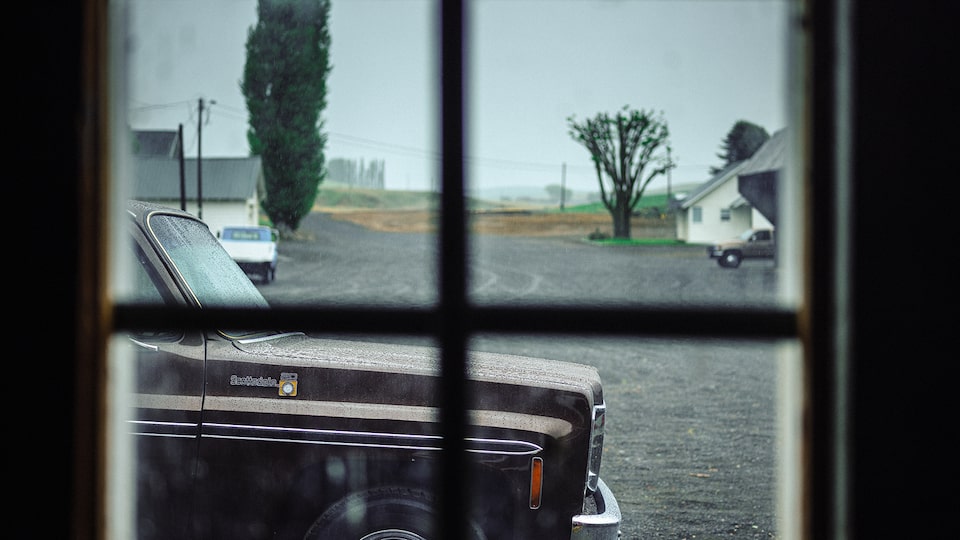 Looking out a 6-paned window of a house onto a gravel lot and the front end of an old Chevy truck.