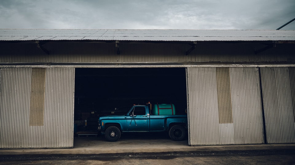 An old blue Chevy pickup is seen through the open doorway of a farm building with a large green tank in the bed.
