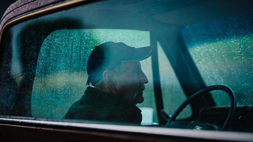 A man sits in the driver’s seat of an old Chevy pickup, seen through a rain-covered window.