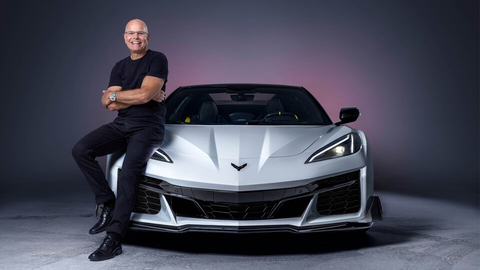 Jordan Lee, with arms crossed and smiling at the camera, leans against the the front right quarter of a white Corvette Z06 that is facing the camera head on.