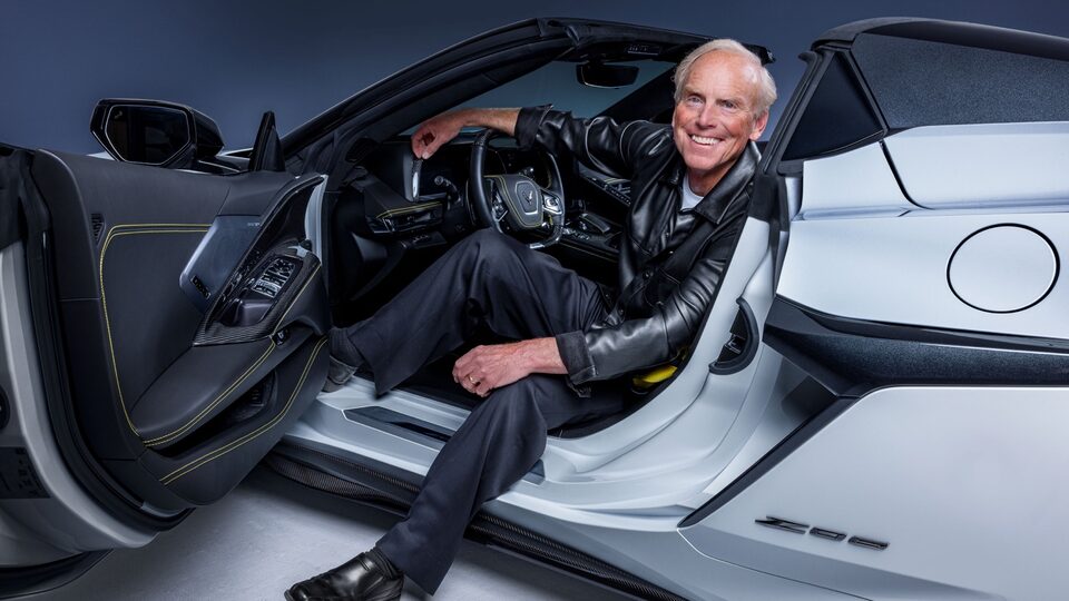 Tadge Juechter smiles at the camera as he sits in the driver’s seat of a white Corvette Z06. The driver’s door is open and he has one foot on the ground outside the car.