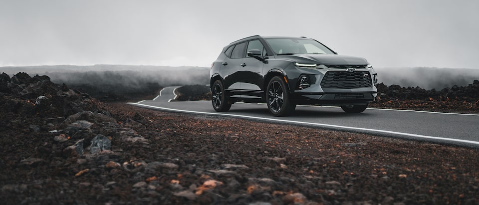 A gray Chevrolet Blazer drives on a narrow strip of pavement through a field of black lava rocks in Hawaii. 