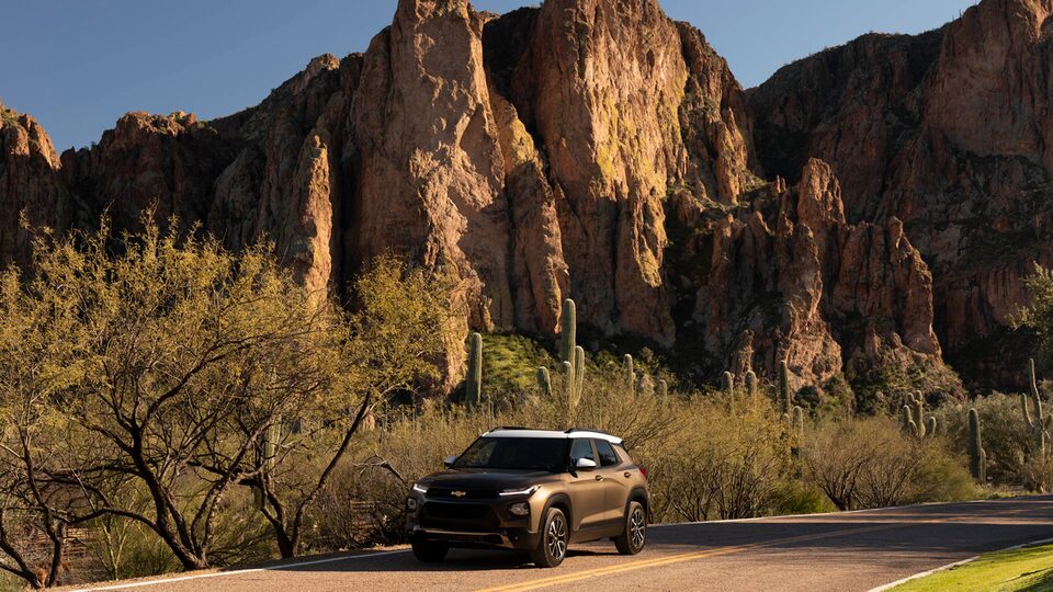 A Zeus Bronze Metallic/Summit White Chevy Trailblazer sits on a dirt road in the desert surrounded by cactus and brush in front of a rocky cliff.