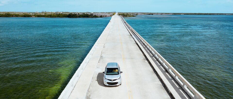 A Silver Ice Metallic Bolt EV drives across a causeway in the Florida Keys, with the ocean on both sides and an island in the background.