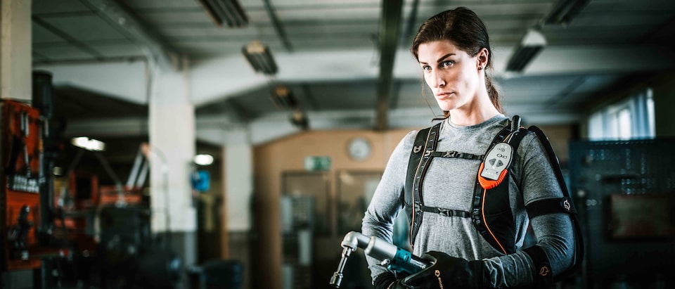 A woman wearing a high-tech, strength-enhancing exoskeleton suit and gloves stands in an industrial plant holding a power wrench.