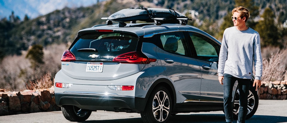 Pro surfer Kevin Schulz walks away from a Bolt EV parked on the side of a mountain road with a surfboard and snowboard strapped to the roof rack on top.