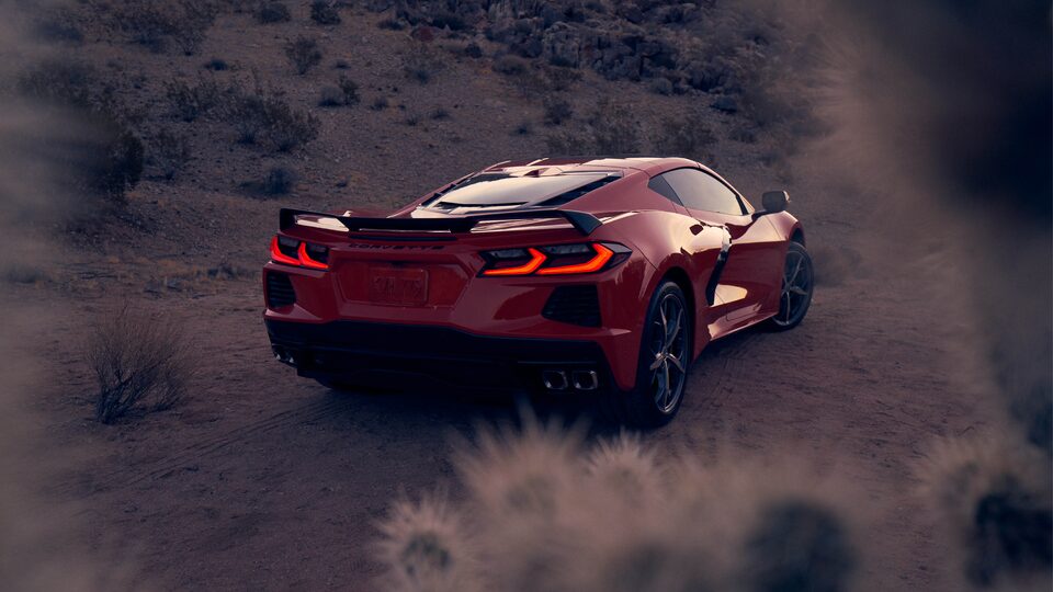 A Torch Red 2020 Corvette coupe in a desert landscape at dusk, seen from the rear with the brake lights illuminated.